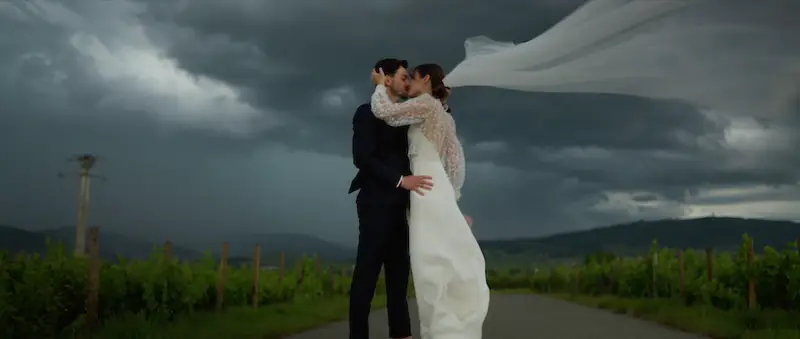Les mariés s'embrassent sur un chemin au milieu des vignes du Beaujolais, sous un ciel d'orage, le voile de la mariée soulevé par le vent