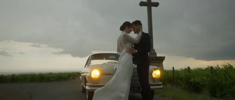 Les mariés enlacés devant une voiture ancienne, près d'une croix au milieu des vignes du Morgon, sous un ciel gris