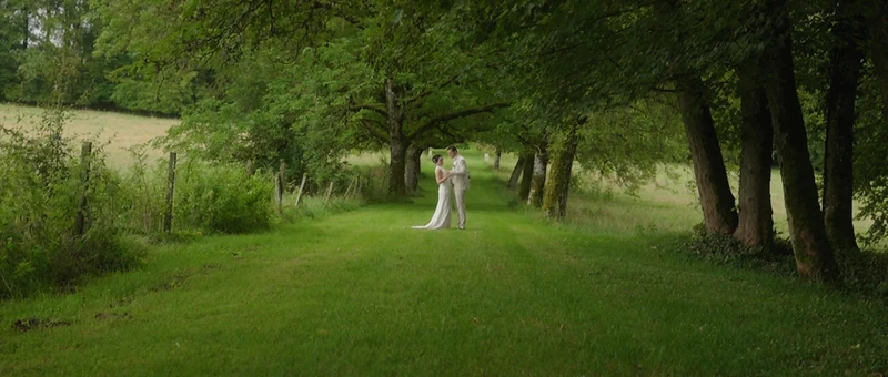 Les mariés au bout d'une allée d'herbe bordée de grands arbres, dans le parc du Domaine du Montremy