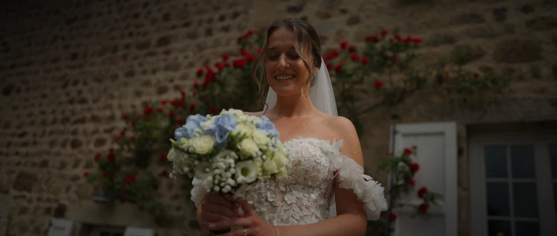 La mariée sourit devant le mur en pierre et les rosiers rouges d'Au Champ Bonheur